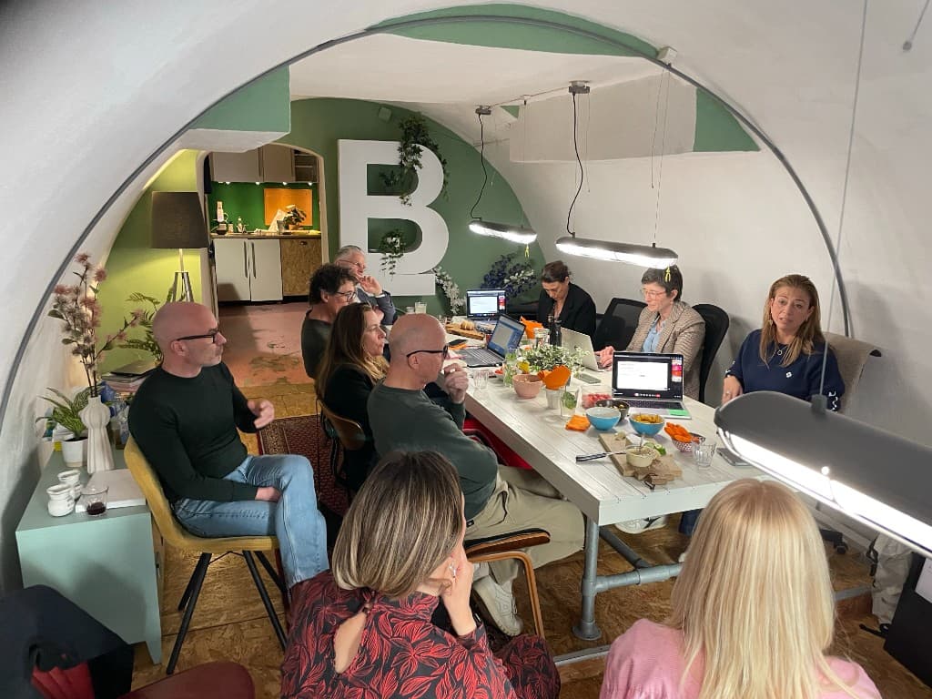 Groep in overleg rond een lange witte tafel in een gewelfde ruimte met groene muur, laptops en snacks op tafel